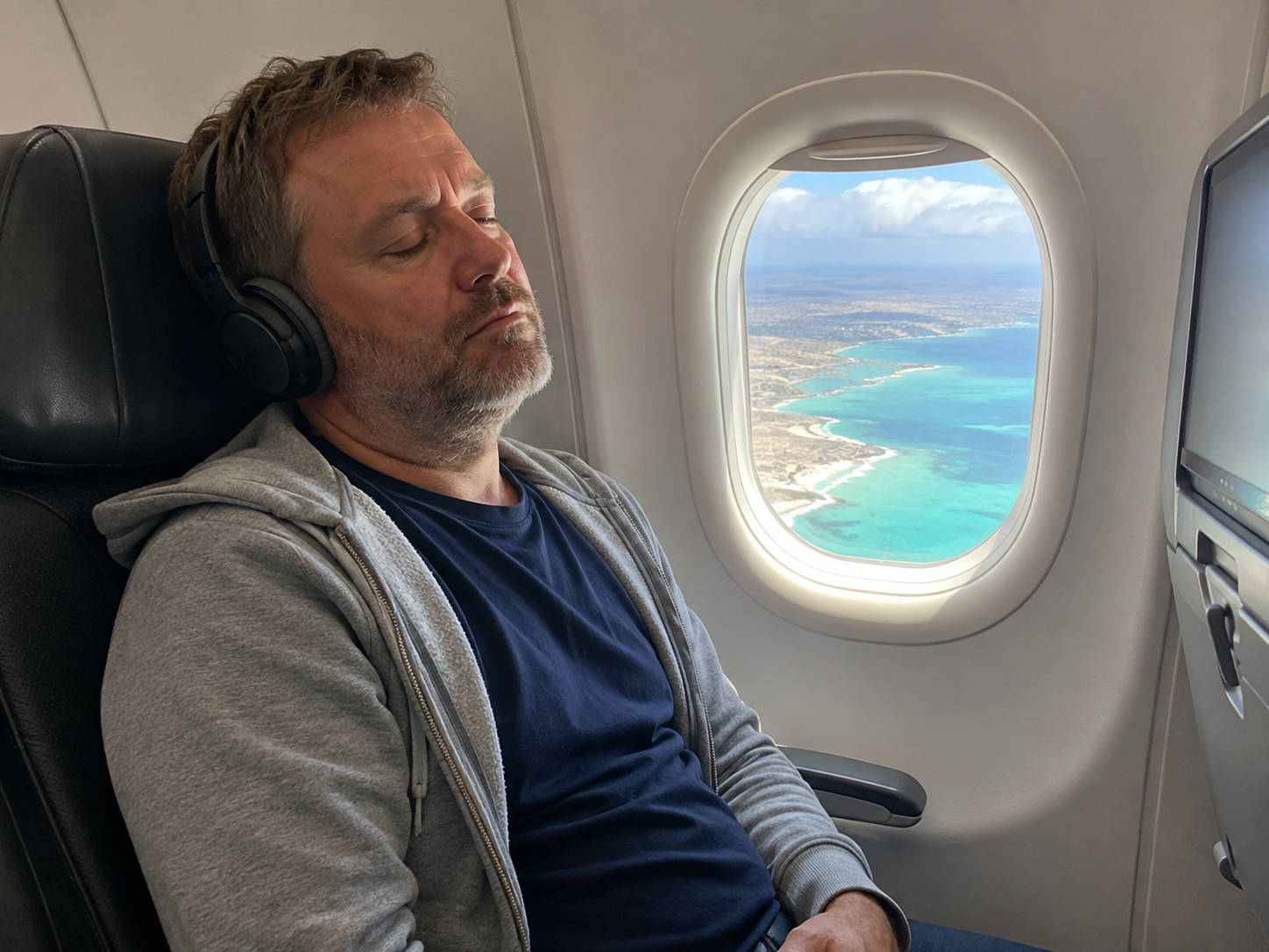 An exhausted passenger rests in a window seat with headphones on, the Caribbean coastline below.